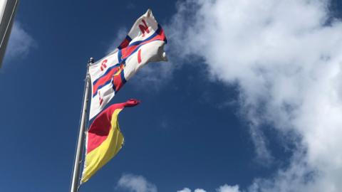 A red and white RNLI flag and a red and yellow flag are blowing in the wind. They are attached to a pole. The sky is blue with a few clouds.