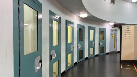 A row of custody suites along a curved wall. The reinforced doors to the custody suites are blue and have lights hanging above them.