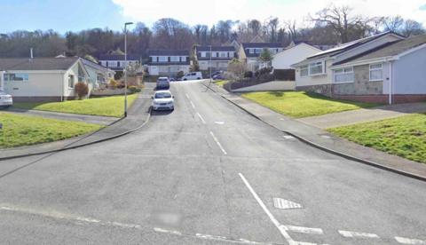 A T junction leading to a road with two cars parked on the left hand side and others in the distance with bungalows on either side of the road and houses at the far end.