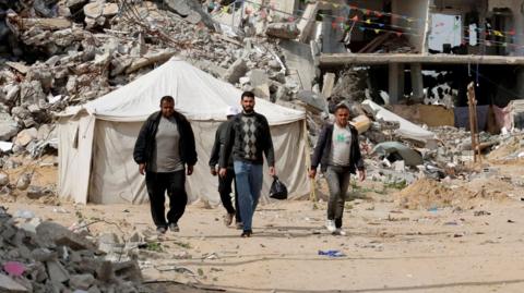 Palestinians walk among rubble in Rafah, with a tent and a bombed out building in the background (March 2025)