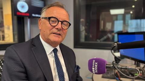 A man wearing a grey suit jacket, white shirt and dark blue tie with white spots on. He is sat in a chair in a radio studio looking into the camera. In front of him is a purple microphone with the words BBC RADIO SUSSEX written on it in white lettering