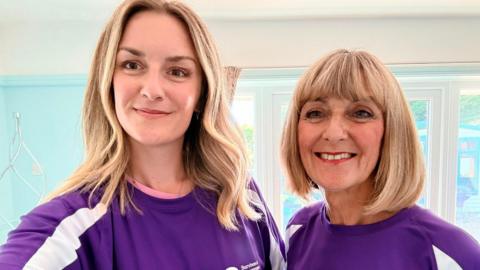 Alyson Noakes and her daughter Siobhan. The pair are wearing matching purple T-shirts and are stood in a house with a blue wall behind them. Alyson has shoulder-length blonde hair and her daughter as slightly longer, wavy blonde hair.