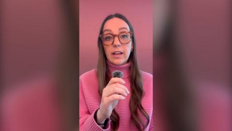 A BBC reporter wearing glasses and a pink jumper holds a mic in a self-shot video