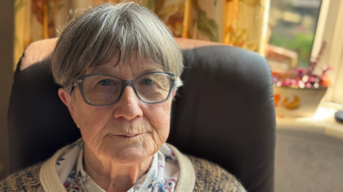 A woman wearing glasses and a white shirt with patterns on it is prominent in the photo. She is sitting in a chair near curtains and a window.