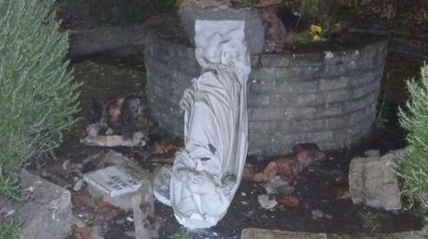 A statue of Our Lady has been toppled off a plinth and partially smashed on the ground. The head has come off. A plaque with writing can be seen broken in half next to the statue.
