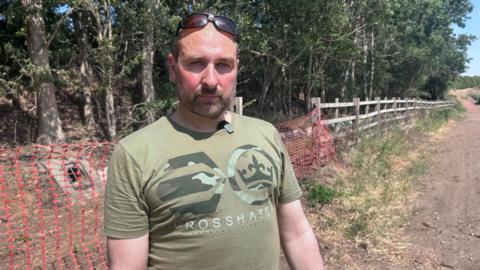 Nick Urquhart is looking at the camera and standing on a mud track. There is a fence and barriers behind him. He is balding with facial hair and is wearing a green T-shirt and has sunglasses balanced on his head.
