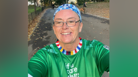 Nikki Gaynor, who is wearing a blue headband and glasses, is taking a selfie while running in a park. She is wearing a green 'if u care share' sports top.