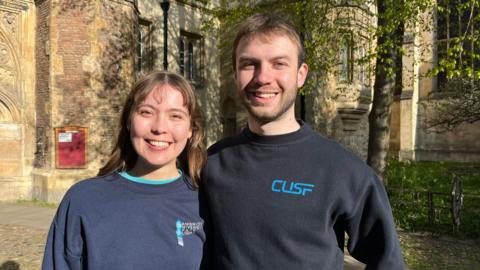 Elisabeth Rakozy and Ben Sutcliffe smiling at the camera in front of Trinity College, Cambridge. They are both wearing Cambridge University Space Flight jumpers. 
