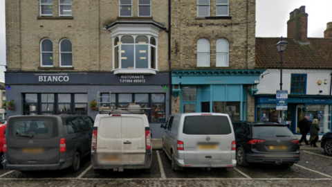 A row of vans and cars parked in front of some shops and restaurants in a market place 