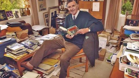 A man in a blue jumper and brown trousers sits on a chair among a pile of books, pictures and clippings and notes