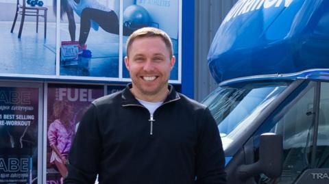 Tom Ryder, who is smiling at the camera, is standing in front of van with Applied Nutrition branding and holding an Applied Nutrition branded water bottle.