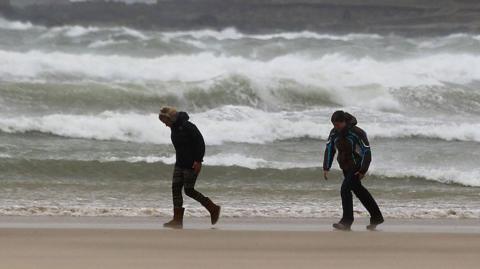 Two people bend into the wind as they walk along a beach with crashing waves behind them