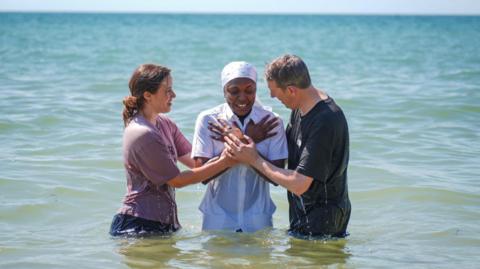 Three people in the sea two either side of a person in all white who has been dunked in the water