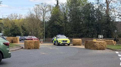 A police car parked on a roundabout which has been blocked by several hay bales that have fallen from a lorry transporting them. Hay can be seen scattered across the road.