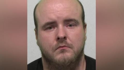 Mugshot of Rogers. He is bald with a scruffy brown beard and left eyebrow piercing.