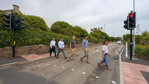 Four men and a woman walk in a line across a road using a puffin crossing with red traffic lights. The road is lined with hedges and there is car in the distance.