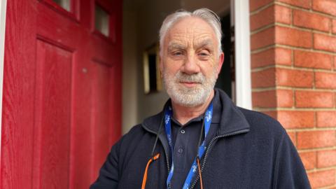 Roger Hewish looking into camera. He has white hair and a white beard, and is standing in front of a red front door which is ajar.