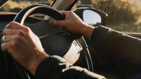 Man holding the steering wheel. He is wearing a black coat and a wedding ring on his left ring finger.