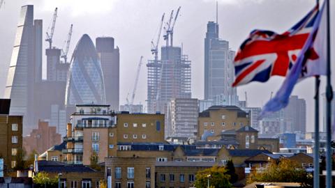 A Union flag flies from a pole near skyscrapers in the City of London