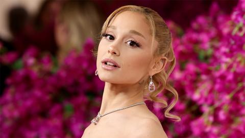 Ariana Grande, a woman looking at the camera with a tilted neck. She is wearing a necklace with the background blurred of pink flowers.