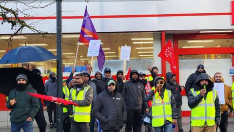 Group of protestors outside an Iceland store in the rain, wearing raincoats, with flags and signs