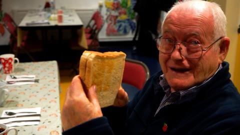 An elderly man smiles as he holds a sandwich
