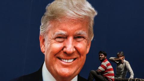 People sit on top of a truck rolling past a poster welcoming US President Donald Trump in Ahmedabad