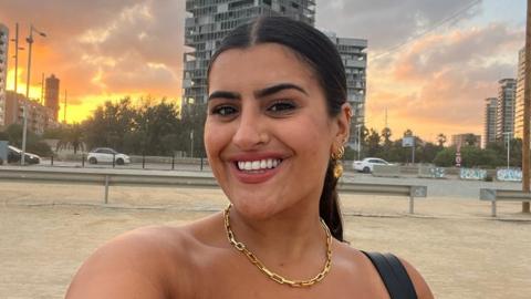 Rosie Breen taking a selfie on a beach. She is smiling and has her brunette hair pulled back in a ponytail. She is wearing a gold chain style necklace and has four gold earrings in her right ear as we look at her. Behind her there is sand and then a road with cars passing by and a single building is directly behind her after the road