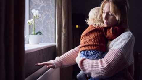 Generic - Mother With Son Trying To Keep Warm By Radiator At Home During Cost Of Living Energy Crisis