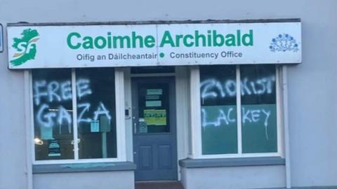 It shows graffiti saying "Free Gaza" and "Zionist Lackey" painted on the windows of a Sinn Féin office in Dungiven