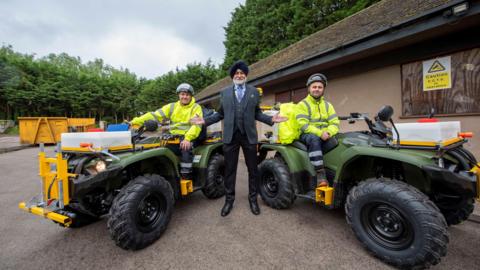 Councillor Bhupinder Gakhal with grounds maintenance team Andrew Harrison Matthew Sheffield. Mr Gakhal is stood in a grey suit between Mr Harrison and Mr Sheffield who are on green quad bikes. 