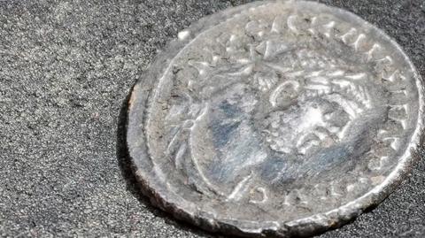 A close-up photograph shows an aged silver coin resting on a dark textured surface. The coin features a worn profile portrait surrounded by an inscription around the edge, although much of the detail has been softened over time. Visible wear and slight damage along the rim suggest the coin has been buried or handled extensively. The image highlights the coin's historical character and archaeological significance.