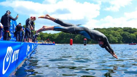 A man wearing a wetsuit dives off platform into a lake.
