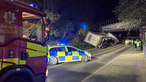 A fire truck, police car and white lorry are visible on the road at night-time. The lorry is leaning at a 45-degree angle at the entrance to the bridge.