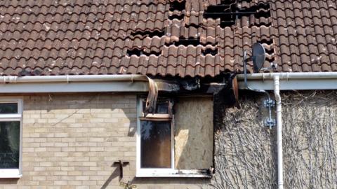 A smoke damaged roof and first floor window. The window is smoke-blackened, and partly boarded up. There are tiles missing from the roof.