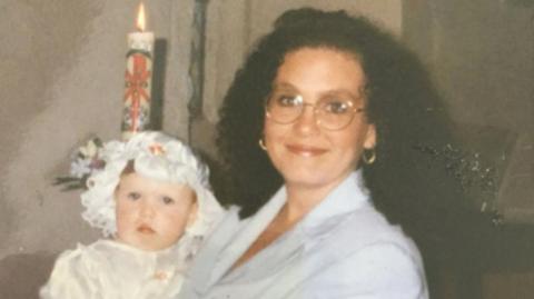 Rochelle Crane, holding her daughter Louise Crane. Rochelle is wearing a light blue shirt and has round spectacles.