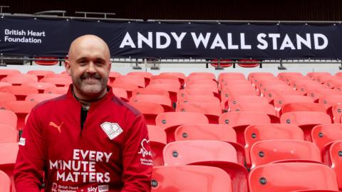 Andy Wall sitting in the Andy Wall Stand at Wembley Stadium. He is looking directly at the camera and smiling and is wearing a red zip-up long-sleeved jacket which says EVERY MINUTE MATTERS on it in white lettering. He is sitting on a red seat. Above him is a sign which says ANDY WALL STAND in white lettering.