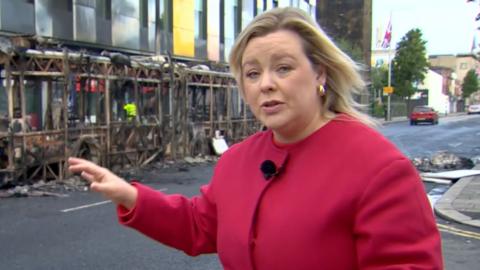 Correspondent wearing red jacket reporting from Belfast, pointing to bus destroyed by fire