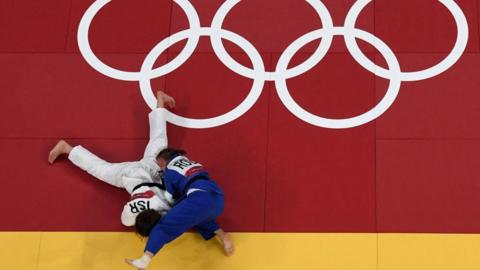 Two judokas wrestle on the floor with five Olympic rings on a red mat
