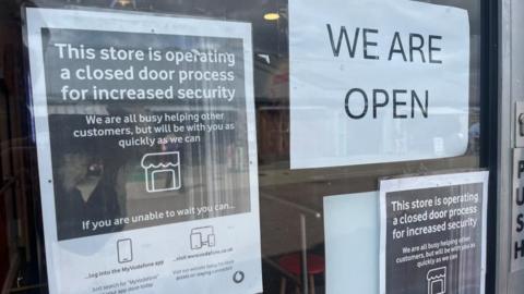 Signs in the Vodafone shop window, stating: "This store is operating a closed door process for increased security. We are all busy helping other customers but will be with you as soon as we can." Another sign states: "We are open".