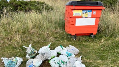 A large orange bin in a grassy field with multiple plastic carrier bags that have the handles tied up on the ground in front of it.