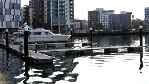 A white boat was in a marina on the left docked up next to a floating pontoon. The pontoons had black tall poles which came out of the water. High rise buildings surrounded the marina.