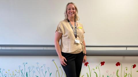 Consultant Nurse Emma Clark stands in the new Peddars Way End of Life Unit at the Queen Elizabeth Hospital in King's Lynn. She is wearing a beige coloured top and black trousers. The bottom half of the walls are subtly painted with daisy's, buttercups and poppies to make the hospital environment seem less clinical. 