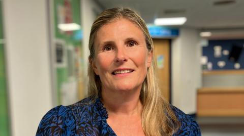 A blonde-haired woman, wearing a blue and black blouse, standing in a gym reception area with a wooden desk and door in the background