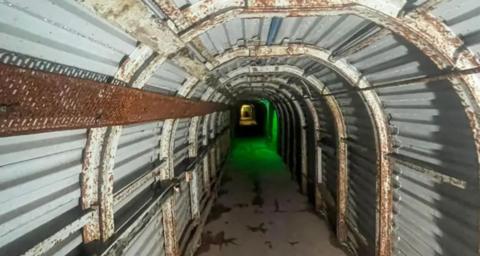 An underground tunnel is lined with corrugated metal and has a concrete floor. An eerie green light can be seen towards the far end