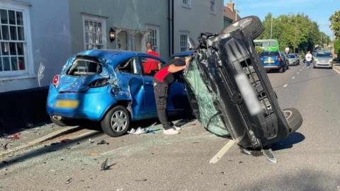 The aftermath of a crash on West Street in Coggeshall. A black car is on its side having turned over. Its windscreen is smashed and there is damage to the front of the car. There is a light blue vehicle which has extensive damage to its rear and side following the crash. There is glass and broken parts of the cars on the ground, and there are two people stood around the crashed vehicles.