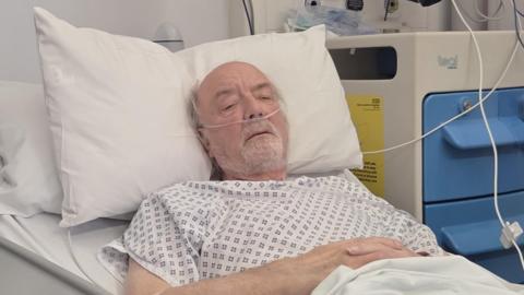John Riding lies poorly in a hospital bed with a tube connected to his nose.