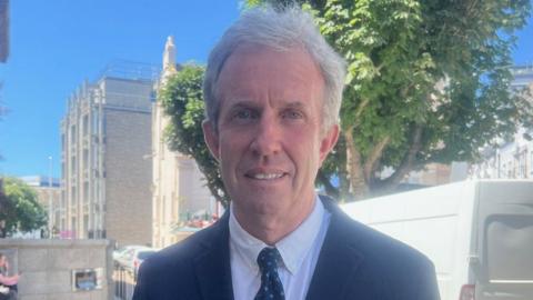 Deputy David Warr is wearing a dark blue suit, white shirt and a black tie with blue patters. He is stood in Jersey's Royal Square. 