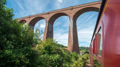 A red passenger train passes beneath a tall brick viaduct with sweeping arches. Lush green foliage surrounds the tracks, contrasting with the warm tones of the masonry. Above, a bright blue sky with wispy clouds frames the scene, highlighting the structure’s scale.
