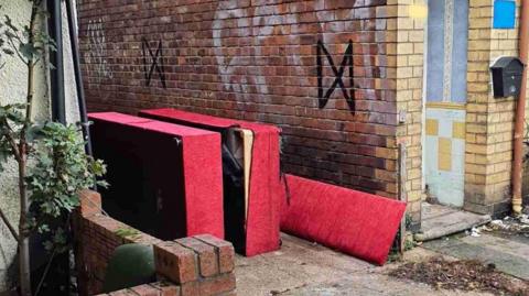 A broken red sofa, which has been dismantled and is upturned on its side, next to a red-brick end-of-terrace house wall that has graffiti on it.